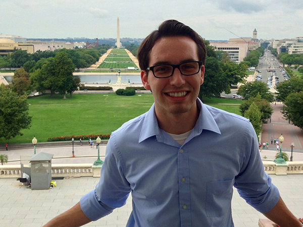 Dan standing in front of the capital building on a sunny day, wearing rectangular black glasses and a light blue button-up. He has a small goatee and his dark hair is combed over to the side.