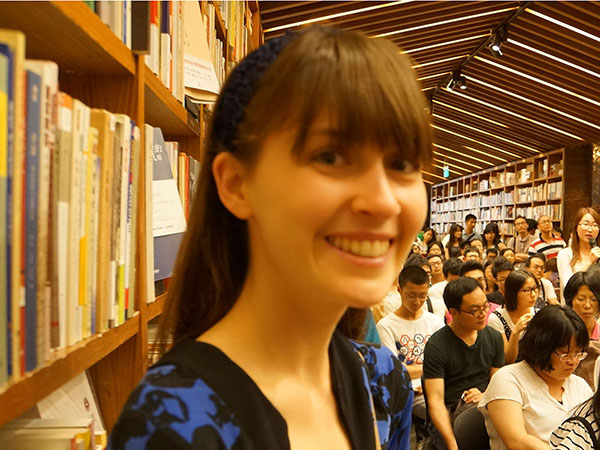 Mika stands in a library surrounded by full bookshelves and dozens of people attending an event. Her brown hair is pulled back by a black headband save for her bangs, and she is wearing a black and blue floral cardigan. 