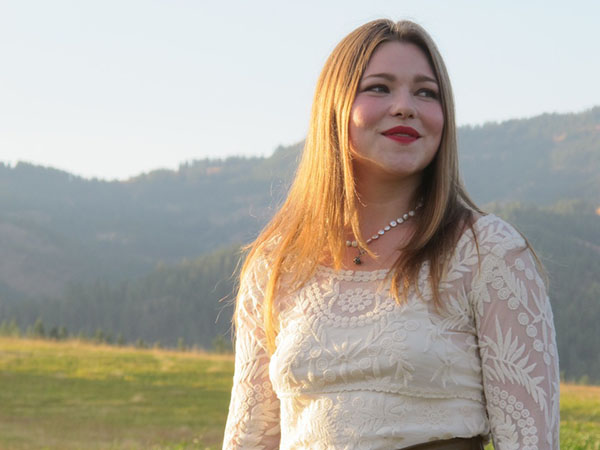 Meredith backlit by the sun in front of a lush green mountain range, meadow, and a reedy pond. She is wearing a white lace dress with long sleeves and a brown decorative belt.