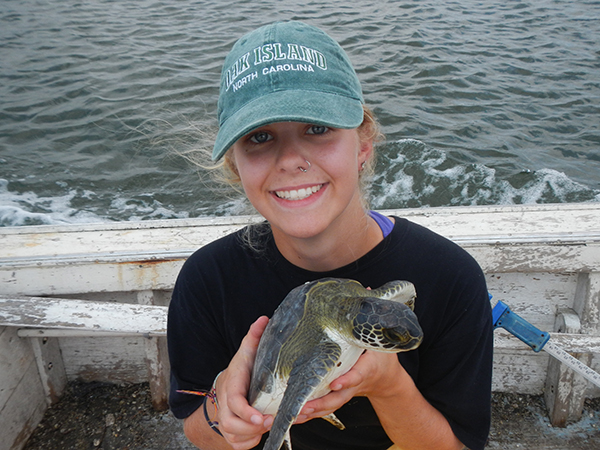 Sarah holds a turtle wearing a black shirt and teal baseball cap while standing on a weathered white wooden boat with the sea behind her.