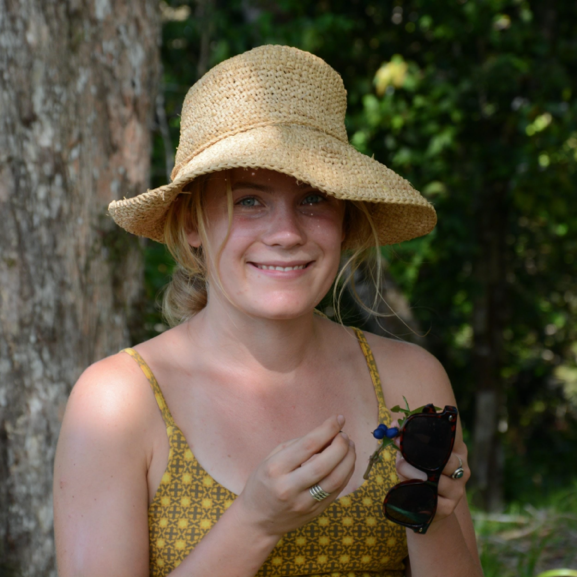 Emily, a smiling blonde, wearing a sunhat and yellow top holds sunglasses and a small blue flower.