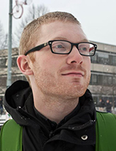 Matthew, a ginger with black rectangular glasses, is wearing a black hoodie under a green backpack and smiling lightly looking out of frame. Behind him it is snowing in the city.