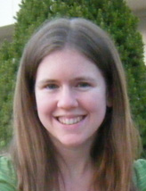 Michaela is smiling at the camera in front of a bush, wearing a green shirt.