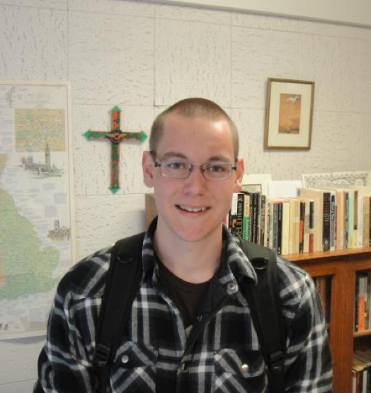 Nathan, a smiling young man with glasses, a black and white plaid flannel, and a shaved head, is standing in front of a wall with a bookshelf, a cross, and a map of some sort.
