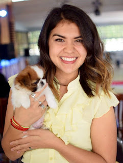 Polly, a smiling brunette with blonde highlights wearing a yellow shirt, holds a small white and brown puppy in her hand.