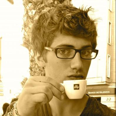 A young Sam in Spain daintily holds a small coffee cup, looking straight at the camera from behind rectangular glasses. His short hair is styled with gel.