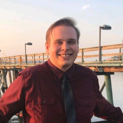 A more recent Sebastian shows him smiling in a dark red button down with a grey tie, standing in front of a pier along the water. 