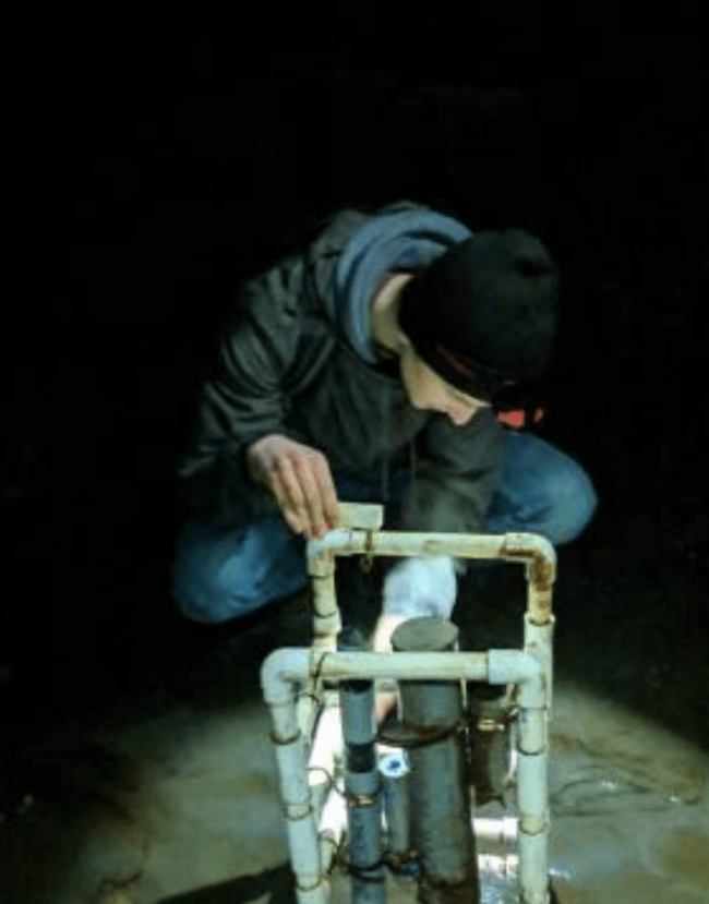 Dexter is wearing a black beanie, a grey hoodie, and blue jeans. He is squatting down, his face obscured by the angle, looking at a contraption of dirty PVC and metal pipes.