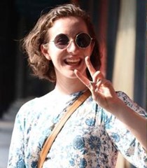A smiling Emmalene is wearing a blue and white floral blouse and round sunglasses, making a peace sign. Her wavy brown hair is cut in a bob.