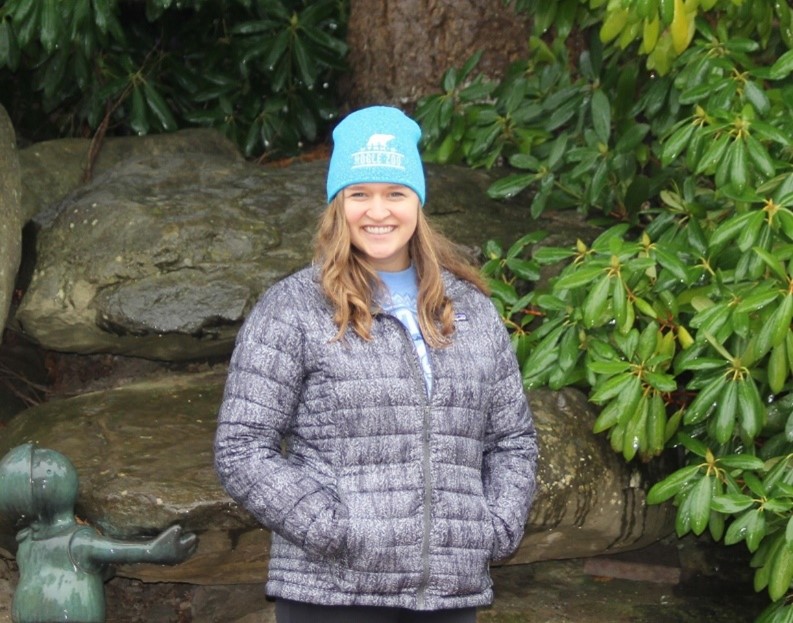 Hillary, a brunette with a bright blue beanie and a light grey down jacket on, smiles at the camera in front of a background of large rocks and bright green rhododendrons. The sculpture of small green men located on Western's campus by the biology building peeks out from the left corner.