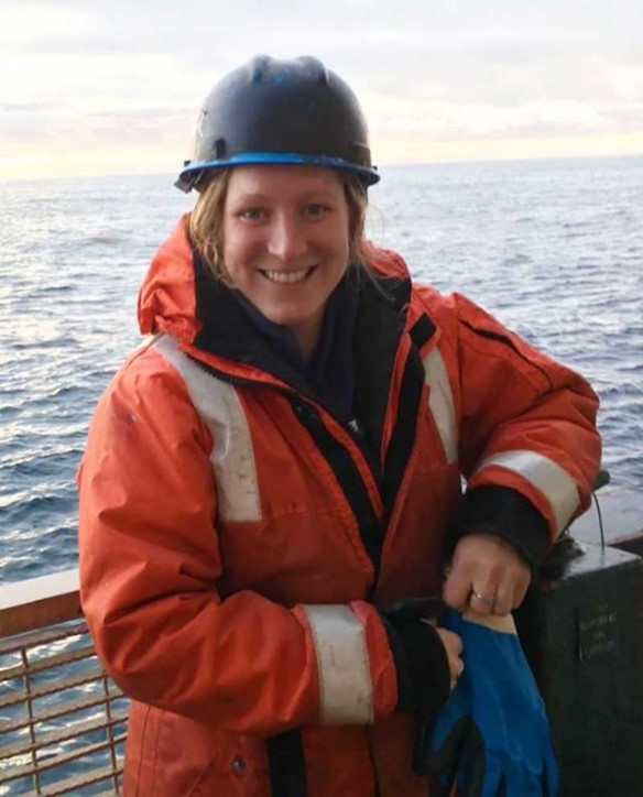 Tanika is smiling, leaning against the rail on a boat with the sea behind her. She has on a blue hard hat, orange reflective jacket, and blue gloves.