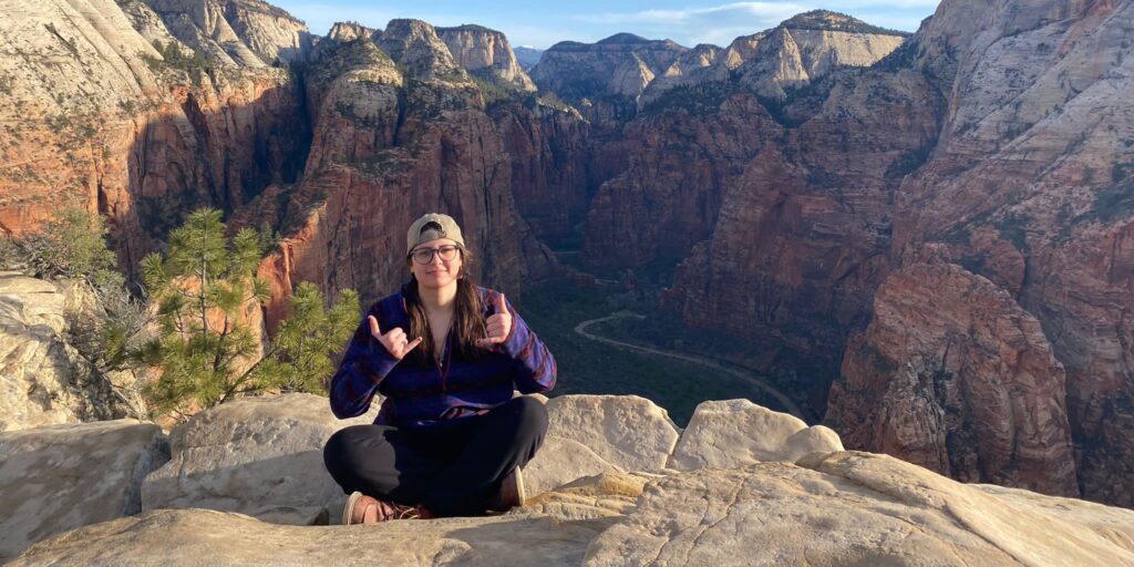 Bobbi sits cross-legged on a rock overlooking a vast desert valley of red rock and minimal vegetation. She is making the 'hang loose' sign with both hands, and is wearing black pants, a maroon and blue flannel shirt, a backwards tan baseball cap, glasses, and tan hiking shoes. 