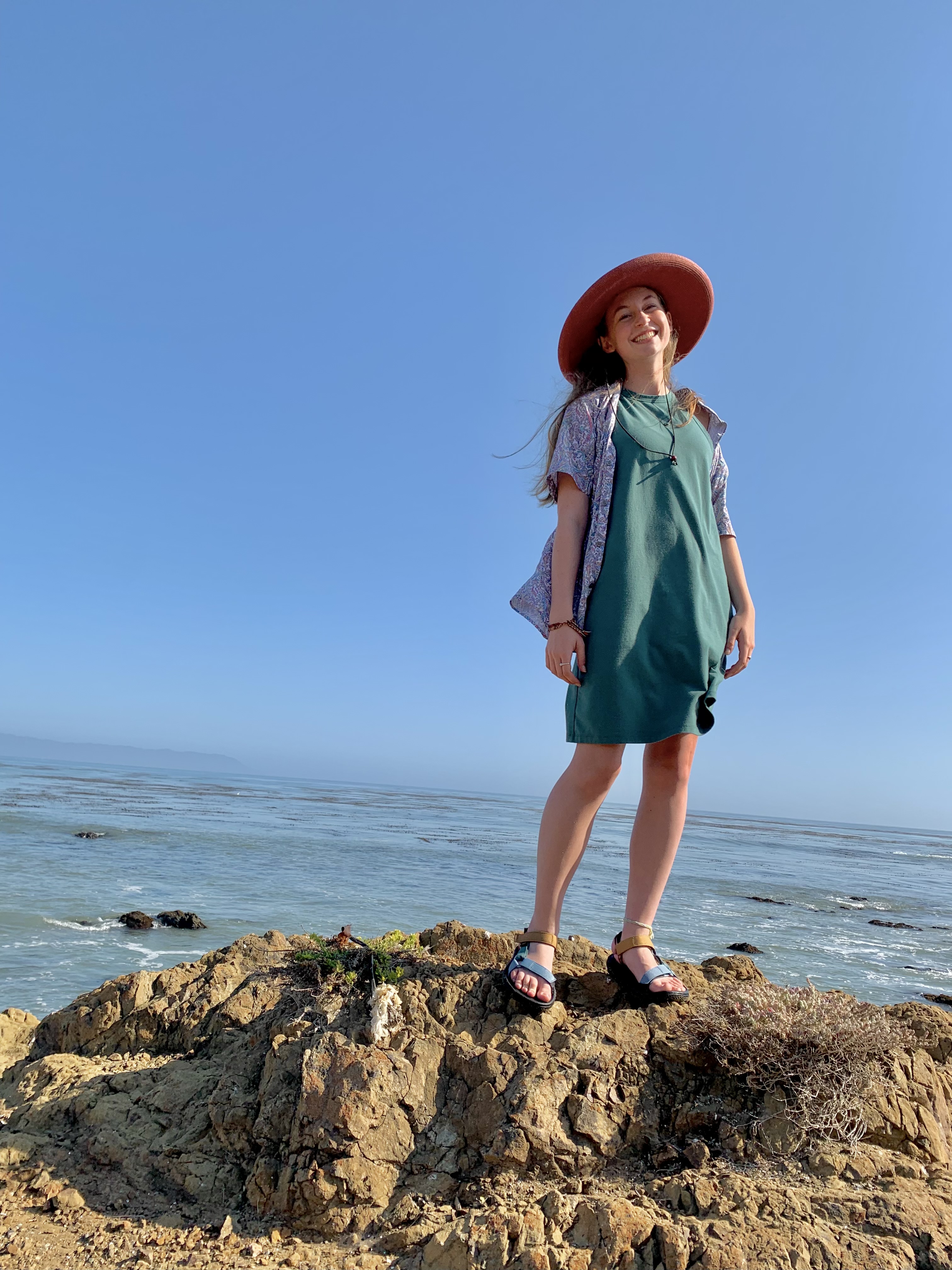 Elise, wearing a teal shift dress under a bright floral button up, stands atop a jagged rock in her sandals. Behind her is the bay and the sky. She wears a burnt orange sun hat over her long, undone brown hair.