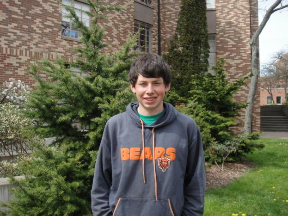 Thomas, a young man with slightly shaggy dark brown hair wearing a grey Bears sports sweatshirt over a green shirt, is standing with his hands in his pockets in front of a brick building, grass, and a couple evergreen trees. 