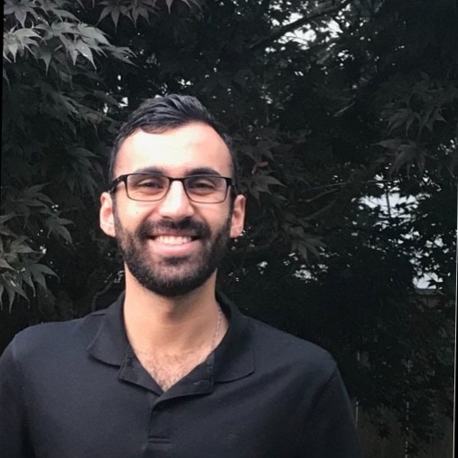 Anton, a young man in a black button up and glasses with short black hair and a short beard, smiles in front of a dark leafy background.