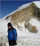Melissa is dressed in a blue parks and a black hat and gloves, standing in front of a snowy mountain peak.