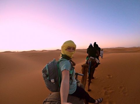 Nicole is wearing a yellow scarf over her head and face, wearing a teal shirt and a grey backpack. She is riding a camel in the desert, with sand dunes below and in front of the camel and a backdrop of a pink and blue sky behind her.