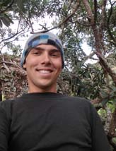 Paul smiles down at the camera wearing a blue checked beanie and a black thermal shirt. Behind him is a tropical forest.
