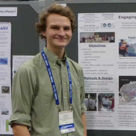 Timothy has shaggy brown curly hair and is wearing a sage green button down with the sleeves rolled up, smiling in front of his scholar's week poster. 