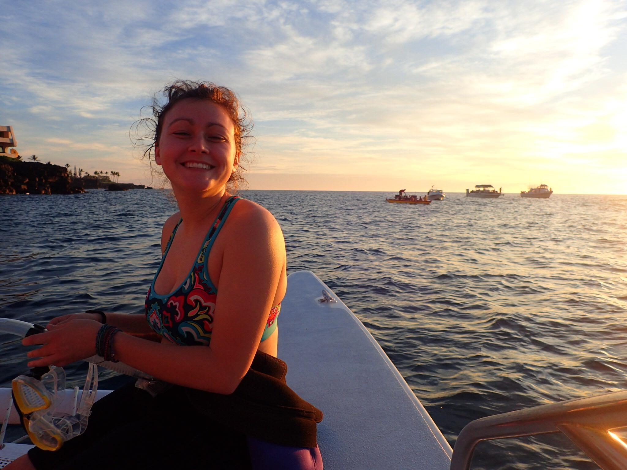 Delaney smiles on a boat holding yellow swim goggles. Her dark air is pulled back and she is wearing a brightly colored patterned swim top. The setting sun casts a glow on her and four boats in the background. 