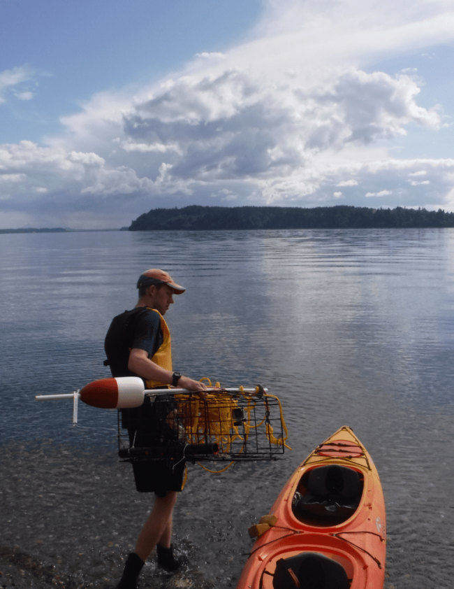 Spencer is dressed in a navy t-shirt, a yellow lifejacket vest, black shorts, black boots, and an orange and grey baseball cap. He is loading a crabbing cage and red and white buoy with yellow rope into an orange kayak. Both he and the kayak are in the shallow end of a vast expanse of water with land in the background some rowable distance away. The sky is blue with cumulous cloud formations.