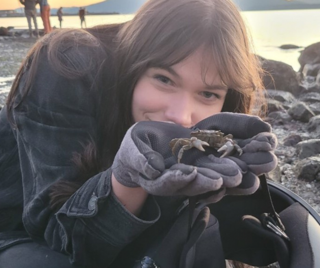 Ula has long dark hair and is wearing a black denim jacket, black leggings, and grey and black gloves. She is crouching and hiding behind her cupped hands holding a startled-looking crab. What looks like a helmet is dangling from her wrist. In the background are assorted rocks on a beach with water and a few people. 