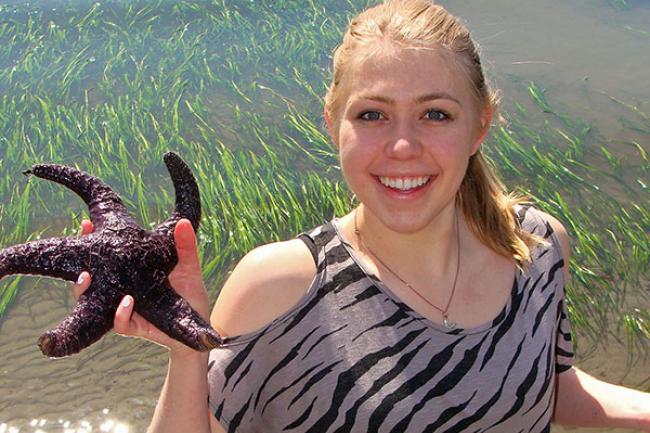 Kaitlyn is smiling standing in the sparkling clear water, holding up a starfish. Her blonde hair is tied back in a ponytail and she is wearing a zebra striped shirt and bootcut blue jeans.