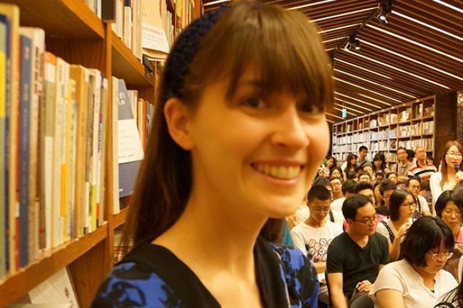 Mika stands in a library surrounded by full bookshelves and dozens of people attending an event. Her brown hair is pulled back by a black headband save for her bangs, and she is wearing a black and blue floral cardigan. 