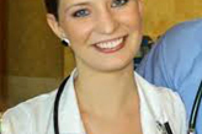 Annaliese is wearing a lab coat with stethoscope around her neck. Her dark brown hair is tightly pulled back, and her dark teal shirt matches her earrings and eyes.