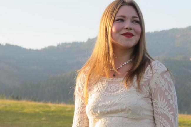 Meredith backlit by the sun in front of a lush green mountain range, meadow, and a reedy pond. She is wearing a white lace dress with long sleeves and a brown decorative belt.