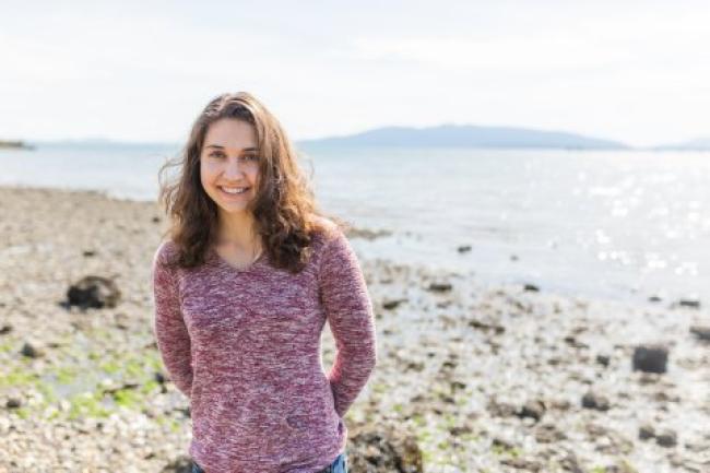 Darby is standing on a rocky beach with the ocean and a faint outline of land in the background. She is wearing a marron-and-white speckled shirt, smiling at the camera,.