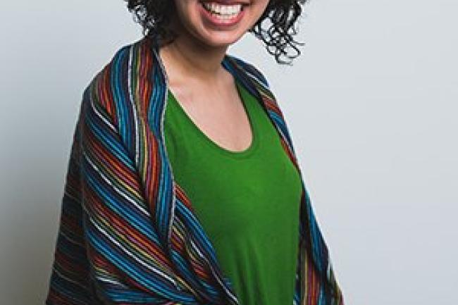 Maria smiles at the camera, wearing a bright green shirt and a multi-colored scarf around her shoulders. She is sitting in a studio with her hands in her lap. 