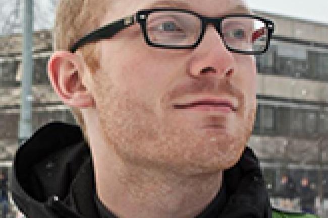 Matthew, a ginger with black rectangular glasses, is wearing a black hoodie under a green backpack and smiling lightly looking out of frame. Behind him it is snowing in the city.
