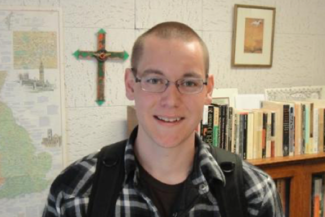 Nathan, a smiling young man with glasses, a black and white plaid flannel, and a shaved head, is standing in front of a wall with a bookshelf, a cross, and a map of some sort.