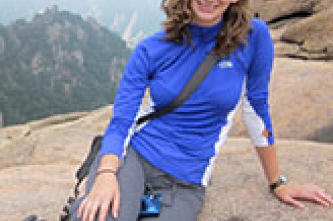 Kelsey is leaning on a boulder above a distant background of a hilly forest obscured by fog. She is wearing a bright blue long sleeve top and grey yoga capris pants with hiking sandals. Her curly brown hair is down and parted on the side.