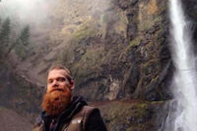 Adam, a redhead with a long beard and his hair pulled back, stands to the left of a tall waterfall in front of a misty cliff. He is wearing a brown down vest with a hoodie underneath. 