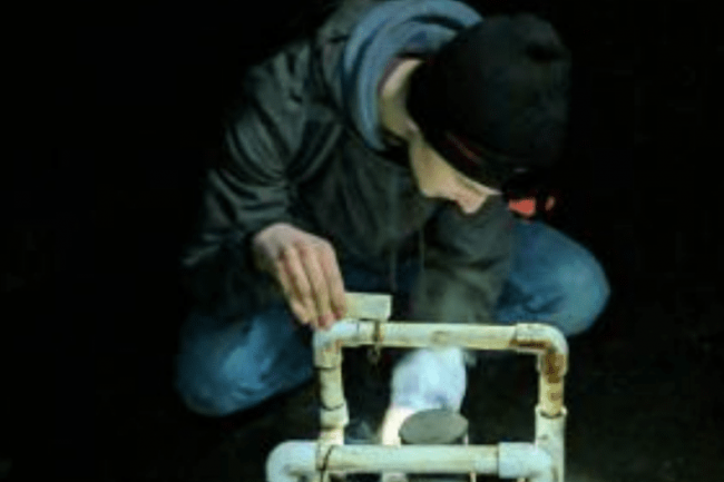 Dexter is wearing a black beanie, a grey hoodie, and blue jeans. He is squatting down, his face obscured by the angle, looking at a contraption of dirty PVC and metal pipes.