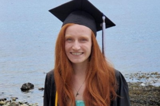 Laura stands smiling in front of the rocky Bellingham bay with the water in the background. Her long bright red hair covers her black graduate robes over an aqua top.