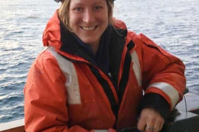 Tanika is smiling, leaning against the rail on a boat with the sea behind her. She has on a blue hard hat, orange reflective jacket, and blue gloves.
