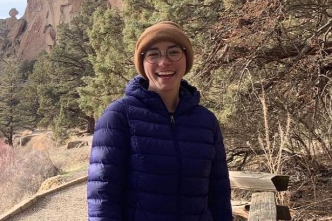 Haley stands in front of a desert landscape with pine trees and high cliffs behind her. She is laughing looking at the camera, and is wearing a tan beanie, black glasses, and a dark blue puffer jacket. 