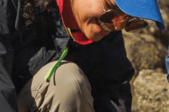 Celida is crouched and smiling at the ground in a rocky area with tidepools. She is wearing a bright blue baseball cap, sunglasses, a black raincoat, tan pants, and black rubber boots. Her long curly black hair is tied back in a ponytail.
