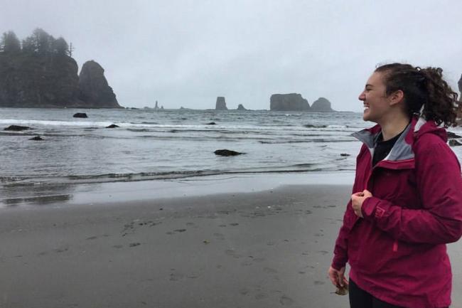 Megan laughs on a coastal beach with sand, water, and rock formations behind her. She is facing left, wearing a magenta rain coat, and her curly brown hair is tied up in a ponytail.