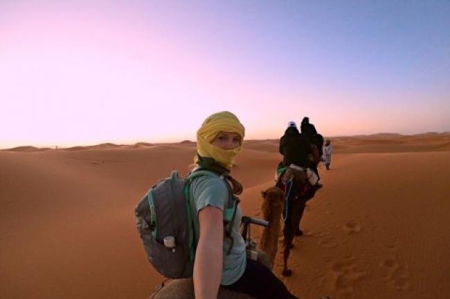 Nicole is wearing a yellow scarf over her head and face, wearing a teal shirt and a grey backpack. She is riding a camel in the desert, with sand dunes below and in front of the camel and a backdrop of a pink and blue sky behind her.
