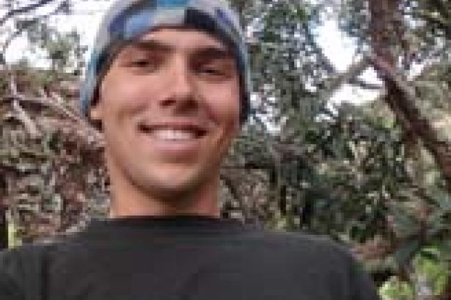 Paul smiles down at the camera wearing a blue checked beanie and a black thermal shirt. Behind him is a tropical forest.
