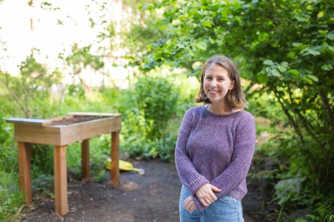 Risa is a smiling woman with shoulder length light brown hair. She is standing in a garden with foliage to the right and a raised gardening bed to the left. She is clasping her hands in front of her, wearing a purple sweater with jeans. 