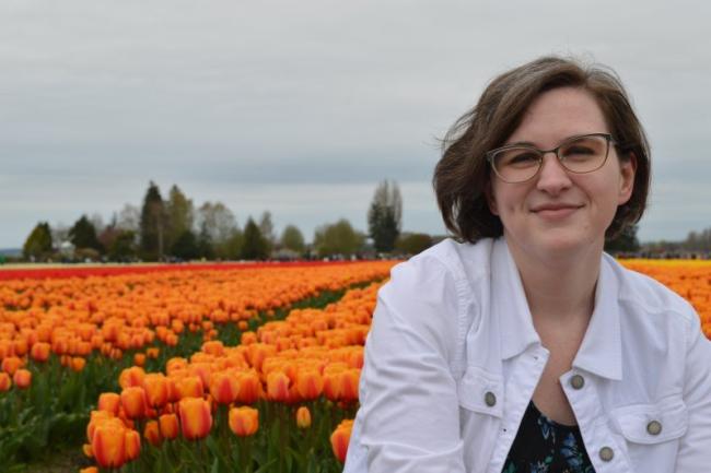 Caitlin is wearing a white denim jacket over a black and blue floral shirt, and has on glasses. Her short wavy brown hair has a streak of grey and is cut in a bob and parted on the side. Behind her is a field of orange tulips in full bloom.