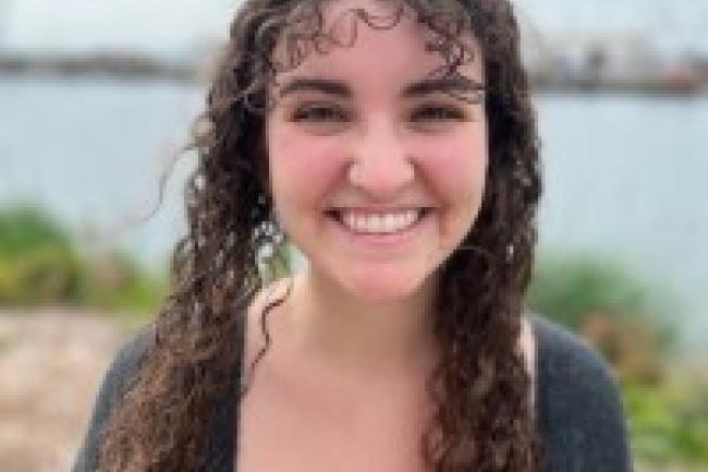 Jasmine smiles in front of a blurry background of the Bay. Her long curly brown hair cover her shoulders, and her bangs are blown sideways by the wind.  She is wearing a dark grey cardigan over a pink and orange striped tank top. 