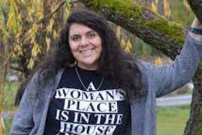 Lindsay smiles at the camera in a grey cardigan over a black and white shirt reading "a woman's place is in the house and the senate." Her long curly and wavy black hair is parted on the side and mostly covers her adorned hoop earrings. She has one arm up leaning against a moss-covered tree branch. 