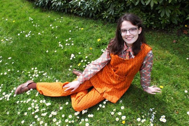 Raven has long dark brown hair and glasses and is smiling sitting sideways on the grass in a patch of daisies. She is wearing a bright burnt orange 60's midi dress over matching pants and a busy brown, red, and black plaid button up. A red and gold tie peeks out from her collar and dress. She has brown socks and brown leather loafers on. 