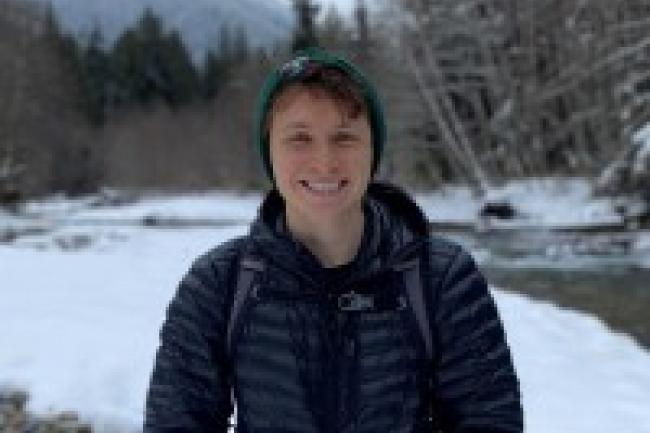 Sam smiles in a green beanie and blue puffer coat in front of a snowy beach with a small amount of rocks and water from the river visible. Behind the stream are snow-covered bare trees, evergreen trees, and a mountain. 